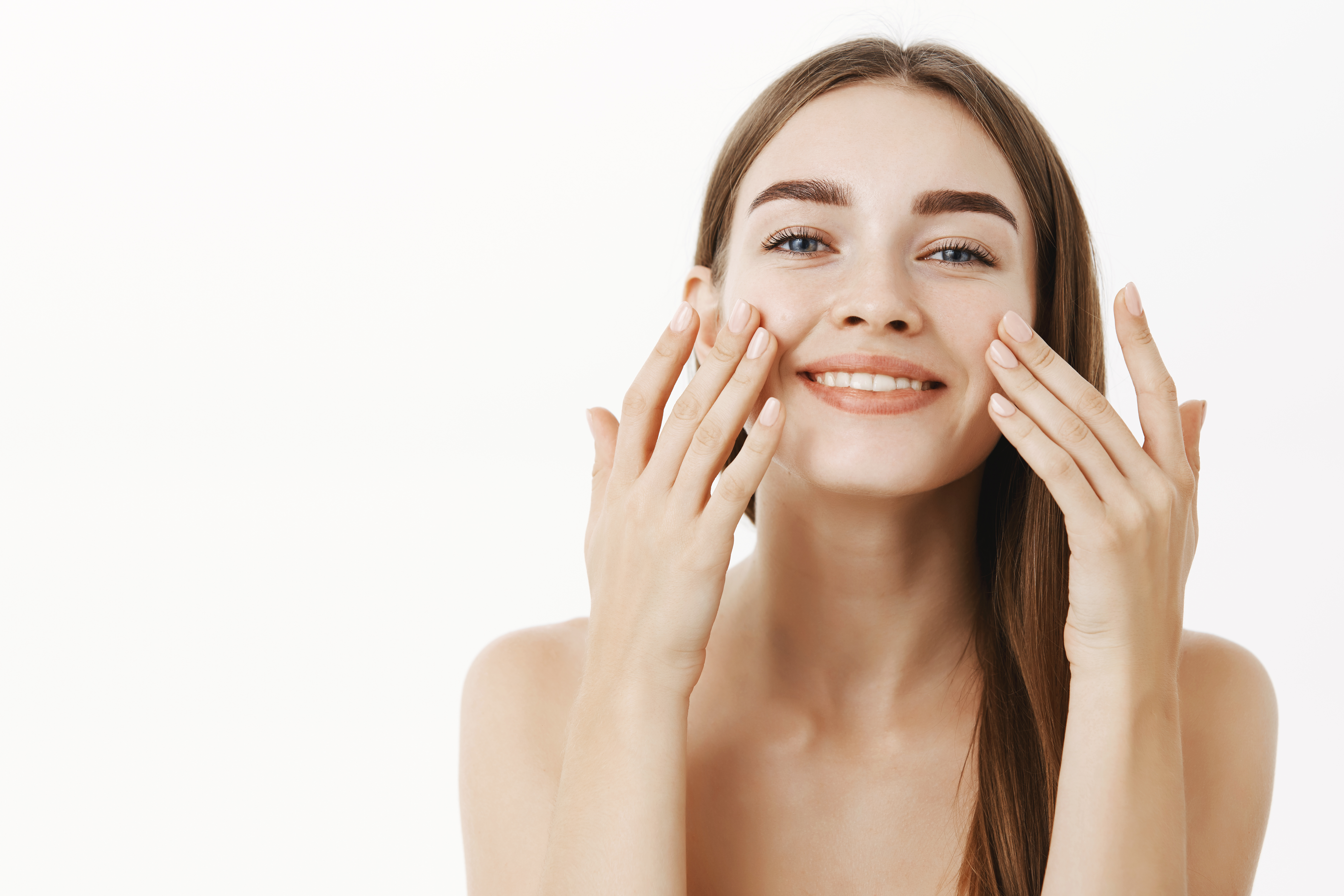 Waist-up shot of charming relaxed and gentle young woman making cosmetological procedure applying facial cream on face with fingers and smiling broadly feeling perfect, taking care of skin.