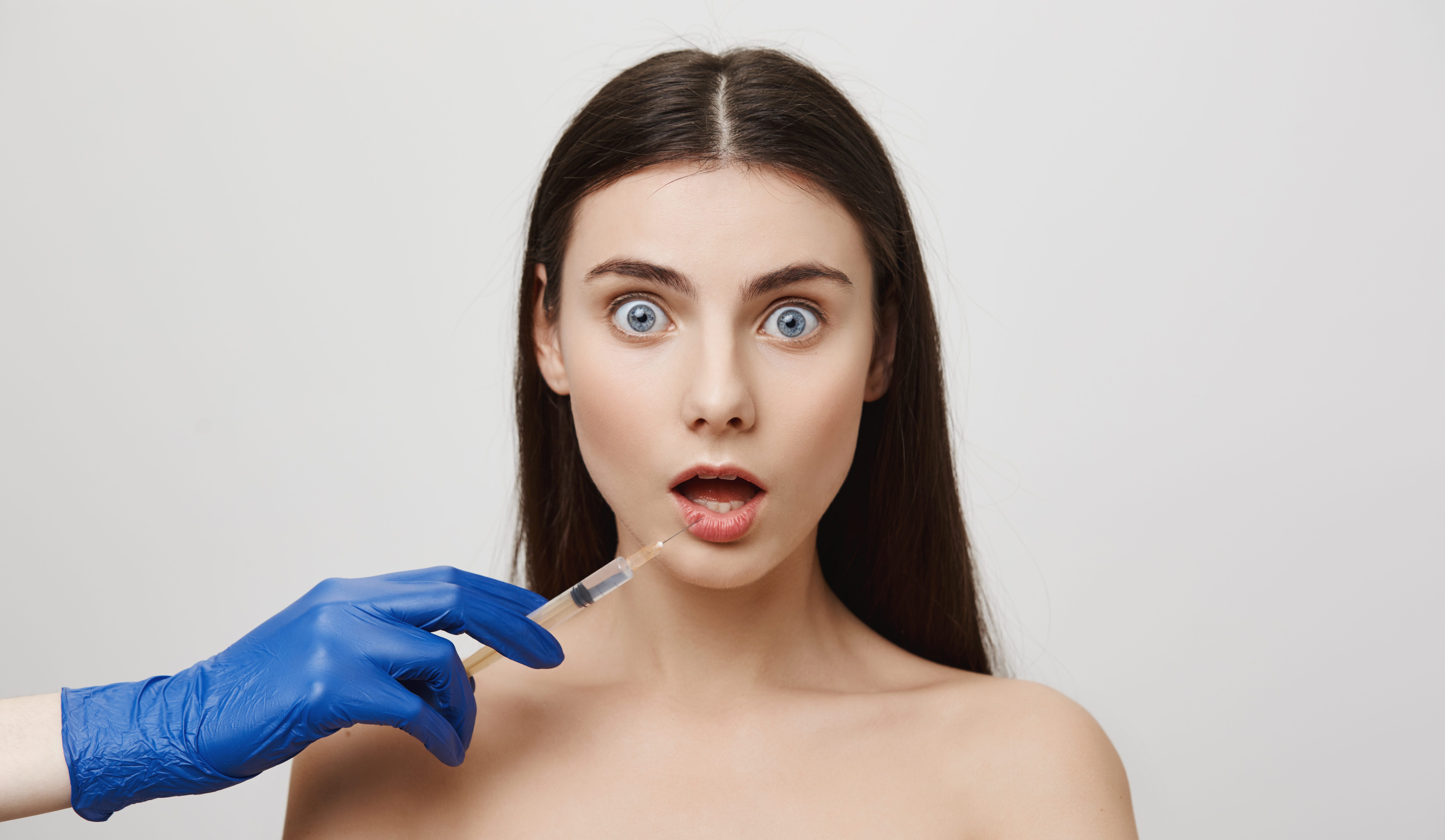 On her way to lip-augmentation. Portrait of nervous young woman receiving shot in lower lip from cosmetologist in medical glove, wanting to change her appearance and make face even more beautiful.
